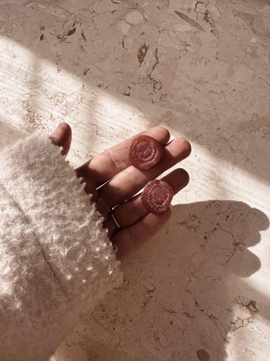 A hand wearing a chunky cream knit sweater holds two small gummies against a textured beige stone background with dramatic sunlight and shadows.