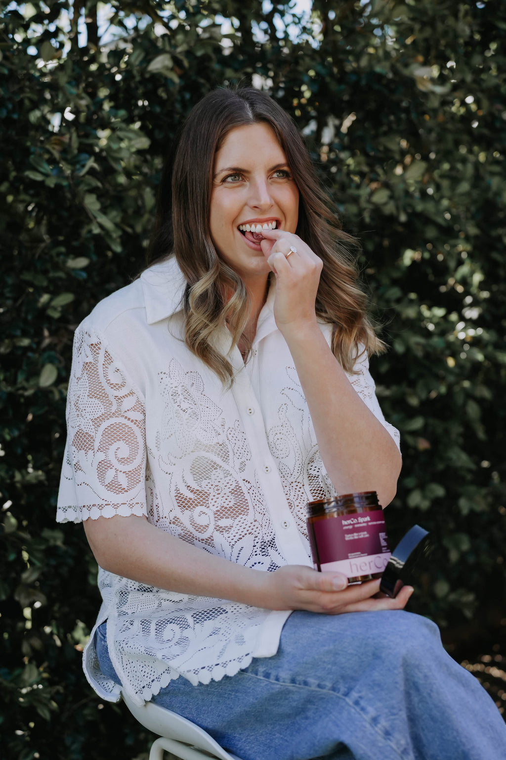 A woman with long brown hair sits outdoors in front of a leafy green hedge, wearing a white lace short-sleeve blouse and blue jeans. She is holding a jar of herCo. Australia gummies product with one hand near her mouth.