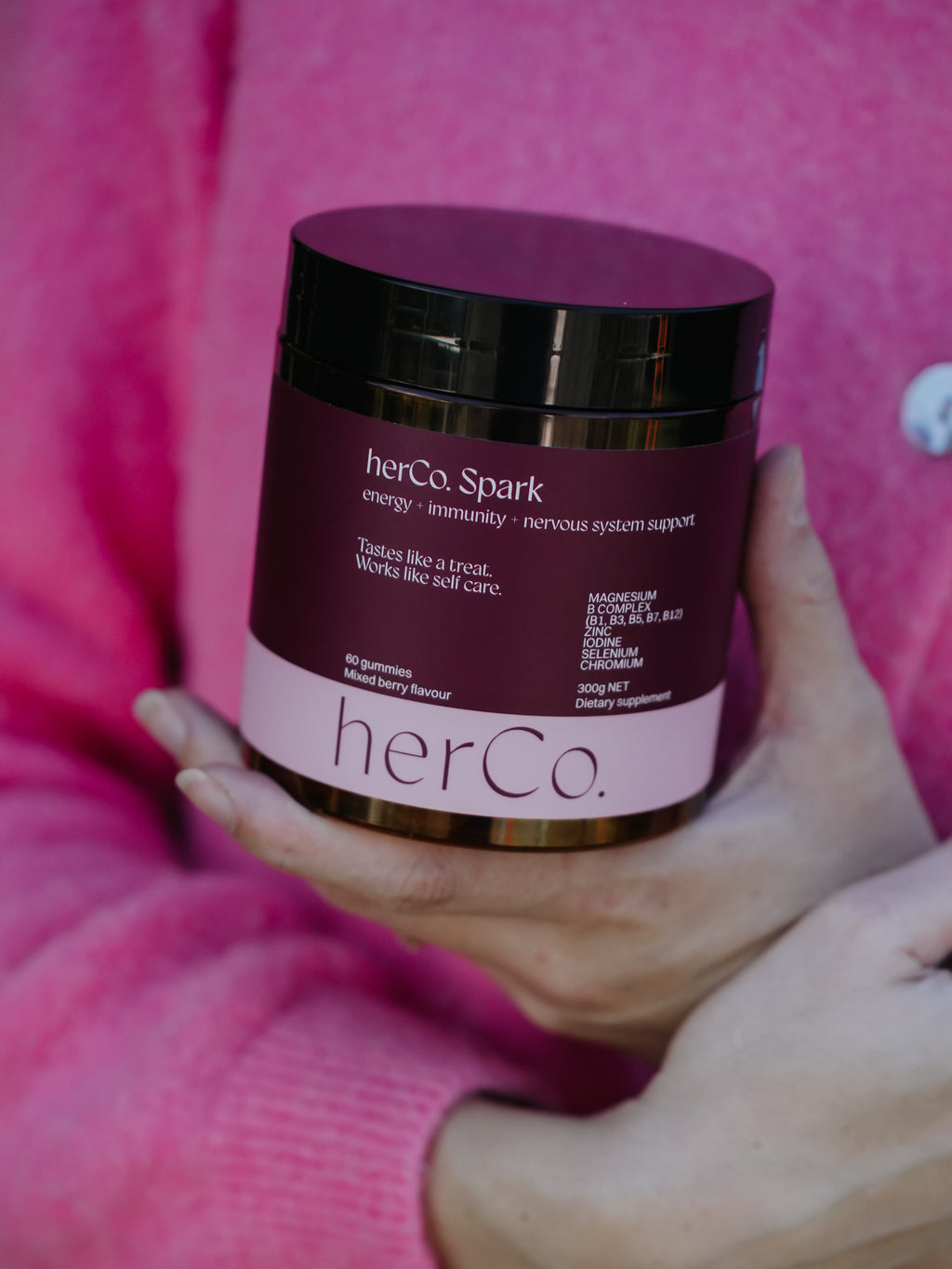 A person in a pink sweater holds a jar of herCo. Spark dietary supplement.