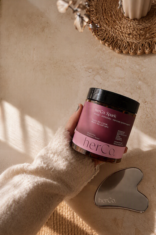 A hand in a cream knit sleeve holds a jar of herCo Spark hair product on a textured beige surface, with a heart-shaped gua sha tool nearby and warm natural light casting shadows.