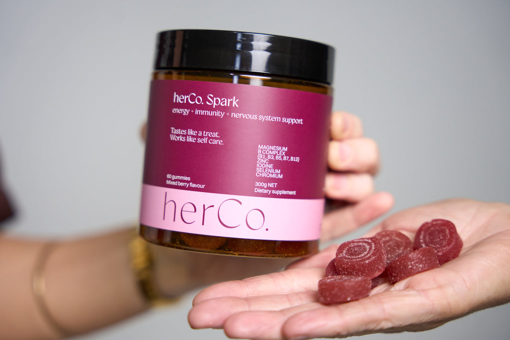 A person holds a jar of herCo. Spark dietary supplement in one hand while holding several pinkish-red gummy candies in the other hand against a neutral gray background.