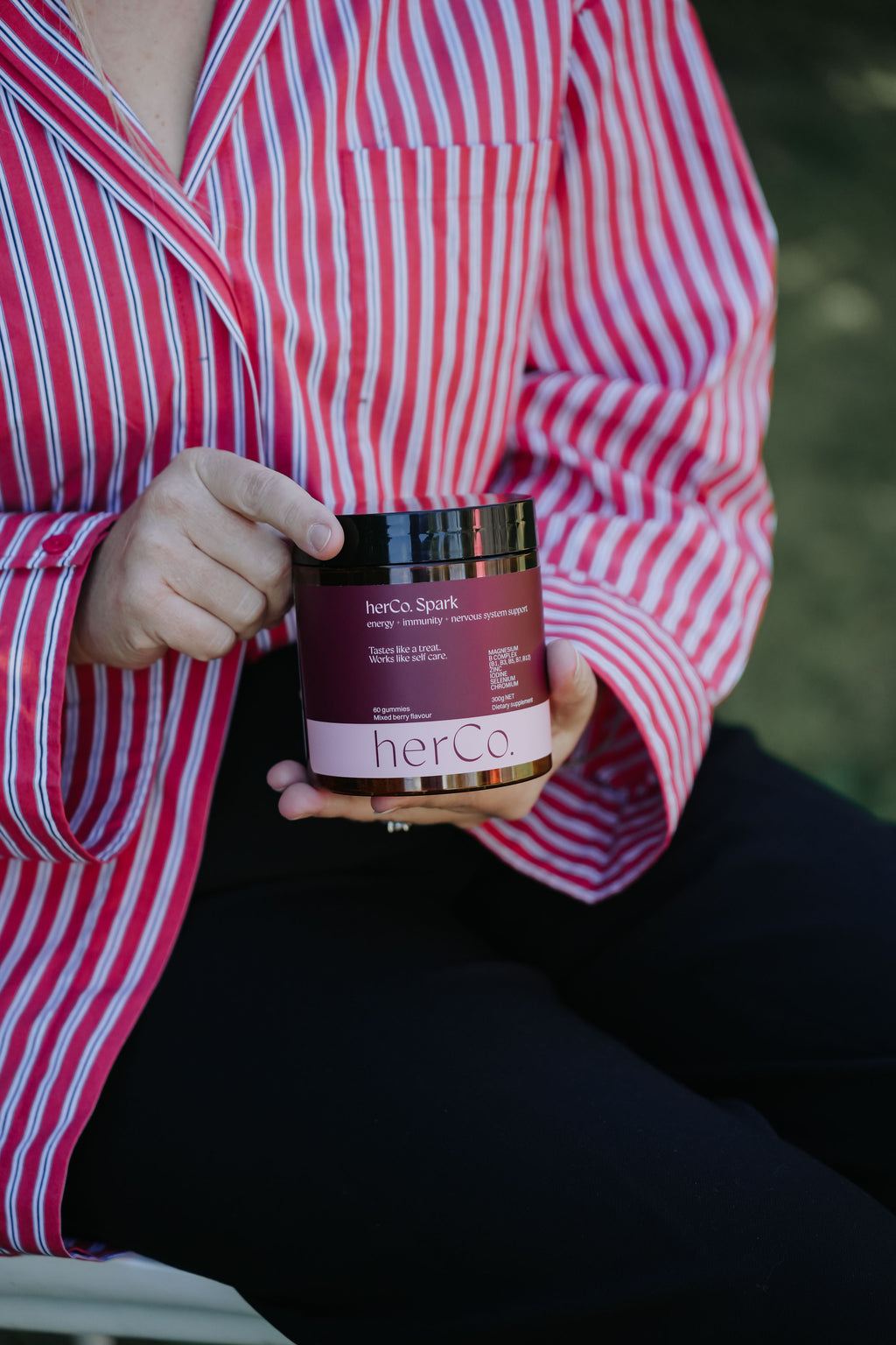 A person in a red and white striped shirt holds a jar of herCo Spark hair product outdoors.